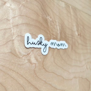 May include: White sticker with black cursive text that reads "husky mom" on a light brown wood background.