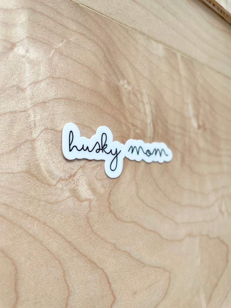 May include: White sticker with black cursive text that reads "husky mom" on a light brown wood background.