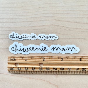 May include: Two white stickers with the text "chiweenie mom" written in black cursive font. The stickers are placed on a wooden surface with a ruler underneath.