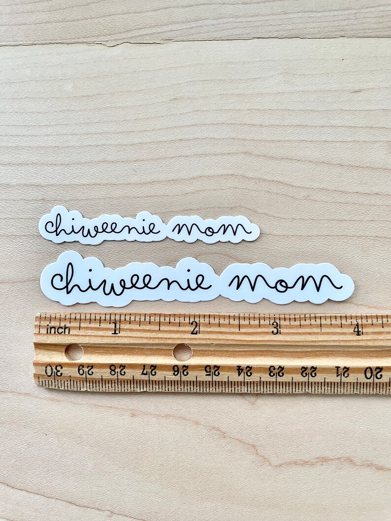May include: Two white stickers with the text "chiweenie mom" written in black cursive font. The stickers are placed on a wooden surface with a ruler underneath.