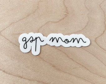 May include: A white sticker with the words "gsp mom" in a black, handwritten font. The sticker is on a light wood surface. The text is in a cursive style.