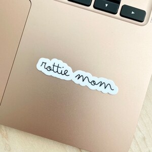 May include: A white sticker with black cursive text that reads "rottie mom" on a gold laptop.
