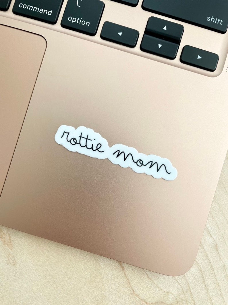 May include: A white sticker with black cursive text that reads "rottie mom" on a gold laptop.
