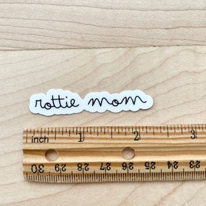 May include: White sticker with the text "rottie mom" in black cursive writing. The sticker is shown on a wooden surface with a ruler for scale.