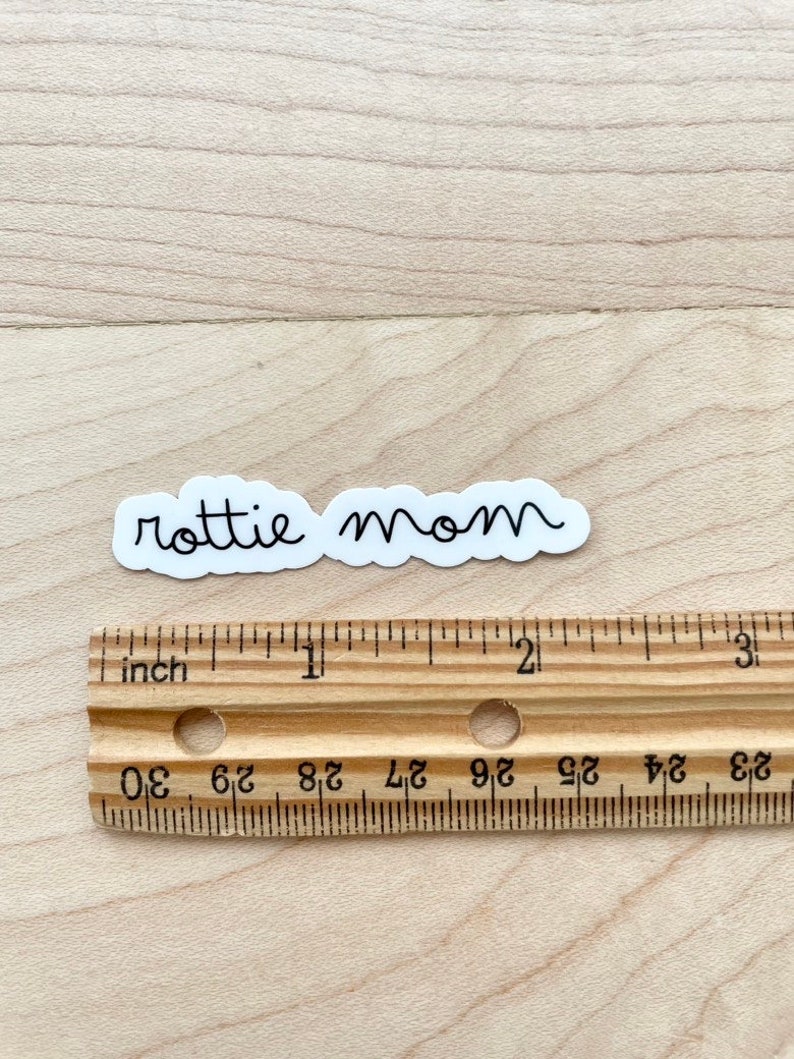 May include: White sticker with the text "rottie mom" in black cursive writing. The sticker is shown on a wooden surface with a ruler for scale.