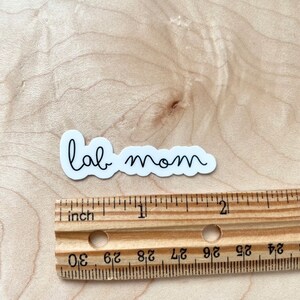 May include: White sticker with black cursive text that reads "lab mom" on a wooden surface with a ruler for scale.