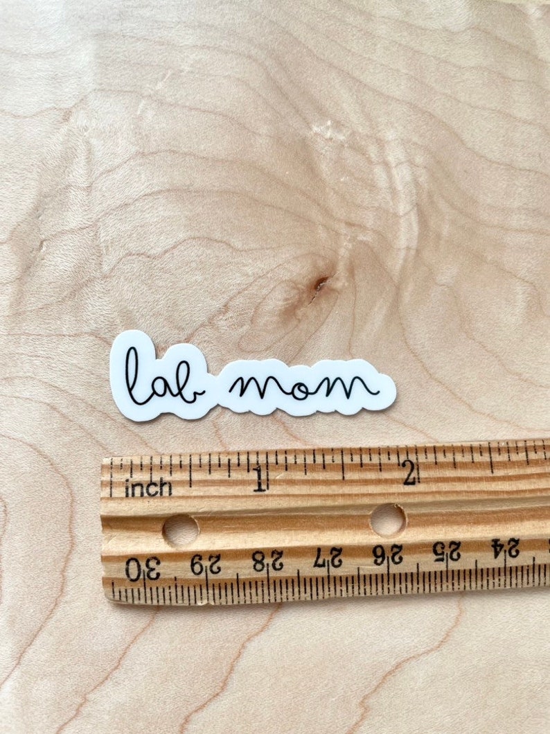 May include: White sticker with black cursive text that reads "lab mom" on a wooden surface with a ruler for scale.
