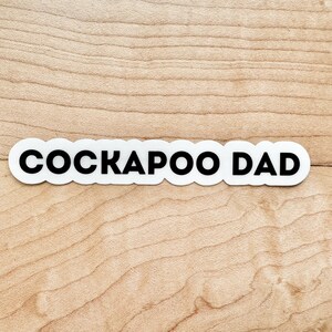 May include: White sticker with black text that reads "Cockapoo Dad" on a light brown wood background.
