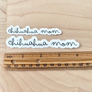 May include: Two white stickers with black text that reads "chihuahua mom". The stickers are on a light brown wooden surface with a ruler.