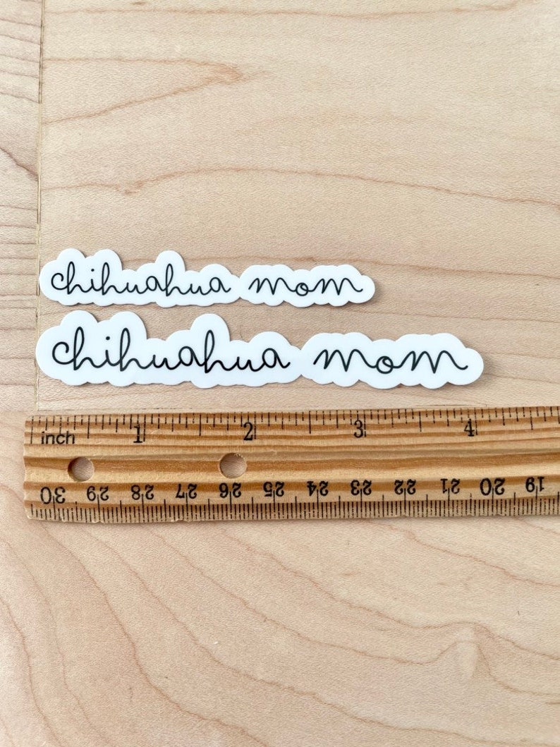 May include: Two white stickers with black text that reads "chihuahua mom". The stickers are on a light brown wooden surface with a ruler.