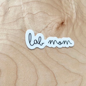 May include: White sticker with black cursive text that reads "lab mom" on a light brown wood background.