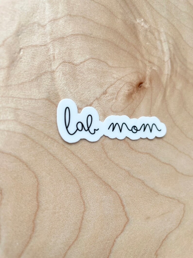 May include: White sticker with black cursive text that reads "lab mom" on a light brown wood background.