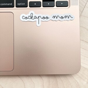 May include: A white sticker with the text "cockapoo mom" written in black cursive script on a rose gold laptop.