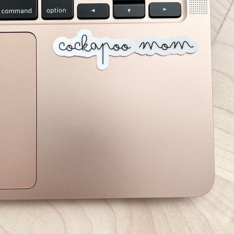 May include: A white sticker with the text "cockapoo mom" written in black cursive script on a rose gold laptop.