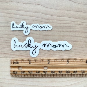 May include: Two white stickers with black text that reads "husky mom" on a wooden surface. A ruler is below the stickers.