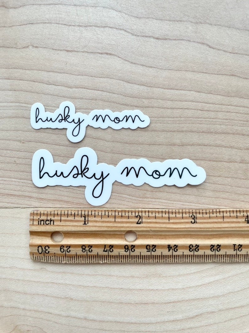 May include: Two white stickers with black text that reads "husky mom" on a wooden surface. A ruler is below the stickers.