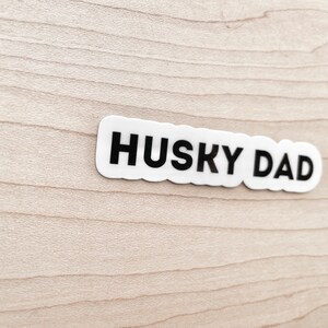 K&ouml;nnte beinhalten: Wei&szlig;er Aufkleber mit schwarzem Text "HUSKY DAD" auf hellem Holzgrund.