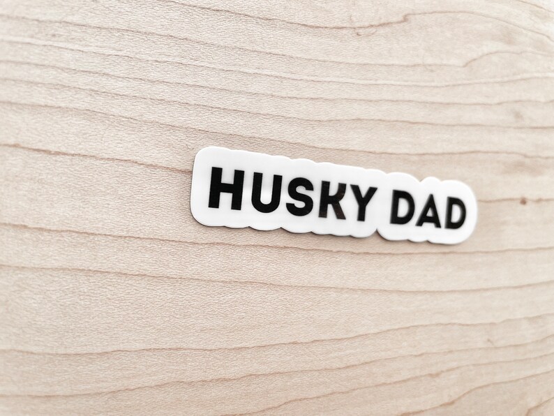 K&ouml;nnte beinhalten: Wei&szlig;er Aufkleber mit schwarzem Text "HUSKY DAD" auf hellem Holzgrund.