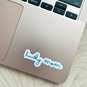 May include: White sticker with black cursive text that reads "husky mom" on a gold laptop.