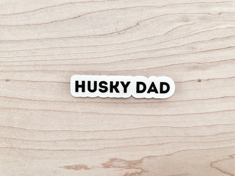 K&ouml;nnte beinhalten: Wei&szlig;er Aufkleber mit schwarzem Text "HUSKY DAD" auf hellem Holzgrund.
