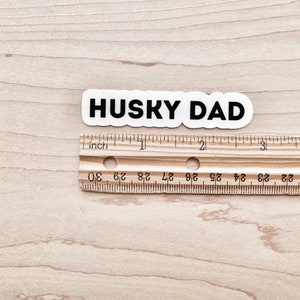 K&ouml;nnte beinhalten: Ein wei&szlig;er Aufkleber mit schwarzem Text, der "HUSKY DAD" lautet, ist &uuml;ber einem Holzlineal platziert. Das Lineal ist in Zoll unterteilt und hat Zahlen von 20 bis 30.