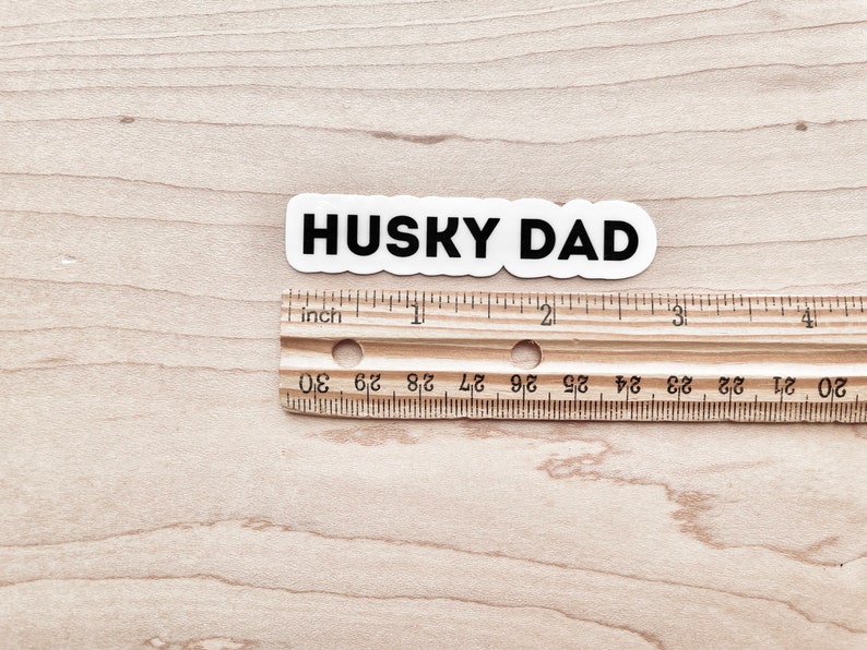 K&ouml;nnte beinhalten: Ein wei&szlig;er Aufkleber mit schwarzem Text, der "HUSKY DAD" lautet, ist &uuml;ber einem Holzlineal platziert. Das Lineal ist in Zoll unterteilt und hat Zahlen von 20 bis 30.