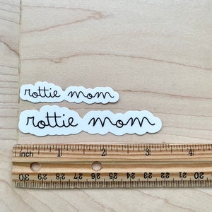 May include: Two white stickers with black text that reads "rottie mom" on a light brown wooden surface. A ruler is below the stickers.