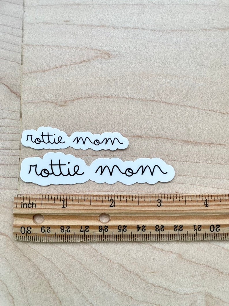 May include: Two white stickers with black text that reads "rottie mom" on a light brown wooden surface. A ruler is below the stickers.