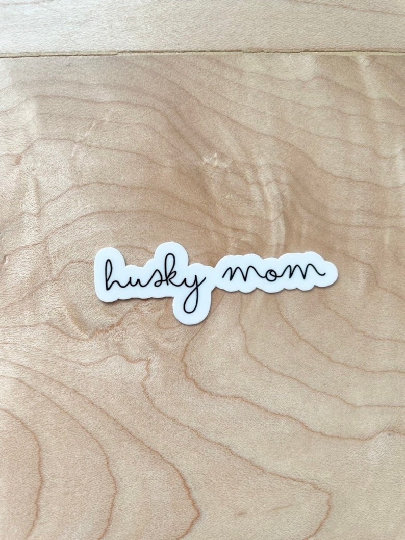 May include: White sticker with black cursive text that reads "husky mom" on a light wood background.