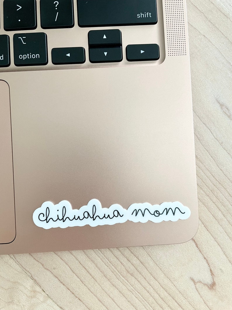 May include: A white sticker with the text "chihuahua mom" in black cursive writing on a gold laptop.