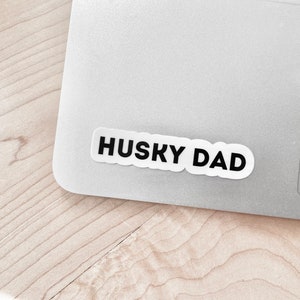 K&ouml;nnte beinhalten: Ein wei&szlig;er Aufkleber mit schwarzem Text, der "HUSKY DAD" lautet, ist auf einem silbernen Laptop angebracht.
