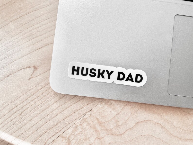 K&ouml;nnte beinhalten: Ein wei&szlig;er Aufkleber mit schwarzem Text, der "HUSKY DAD" lautet, ist auf einem silbernen Laptop angebracht.