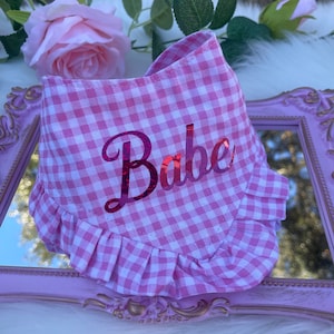 May include: Pink and white gingham dog bandana with a pink ruffled edge and the word "Babe" in pink glitter lettering.