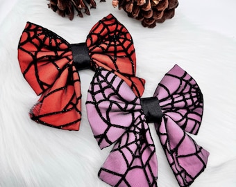 May include: Two Halloween-themed hair bows. One is orange, the other is purple, both with a black spiderweb design. The bows are made of fabric and have a black satin center. The bows are displayed on a white, fluffy surface.