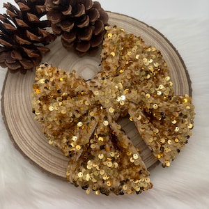 Può includere: Un grande fiocco dorato con paillettes, probabilmente per accessori per capelli, è appoggiato su una fetta di legno. Il fiocco è ricoperto di piccole paillettes lucenti. Due pigne sono sullo sfondo. Un accessorio per capelli festivo.