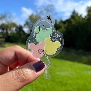 May include: A clear sticker with three balloons in the shape of Mickey Mouse ears. The balloons are green, yellow, and pink. The sticker has the text "You can fly" written on it.