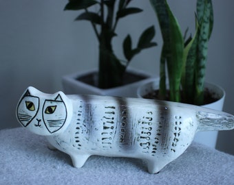 Może przedstawiać: Biała ceramiczna figurka kota z czarnymi i żółtymi detalami malowanymi. Kot leży z głową odwróconą w bok i ogonem zwiniętym. Figurka ma fakturowaną powierzchnię.