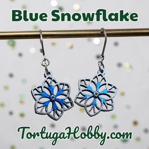 May include: A pair of silver snowflake earrings with blue accents. The earrings are hanging from silver hooks. The text "Blue Snowflake" and "TortugaHobby.com" are displayed in the image.