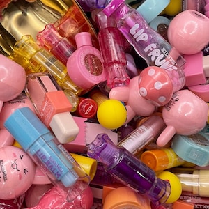 Op de afbeelding: Een verscheidenheid aan kleurrijke lippenbalsems en lipgloss in verschillende vormen en maten. Sommige van de lippenbalsems zijn in de vorm van een vis, een paddenstoel en een hart. De lippenbalsems zijn in verschillende kleuren verkrijgbaar, waaronder roze, rood, oranje, geel en paars. De lippenbalsems zijn in een stapel gerangschikt, waarbij sommige elkaar overlappen. Alle lippenbalsems bevinden zich in doorzichtige plastic verpakkingen, en sommige hebben etiketten met de woorden "Lip Balm" en "Lip Fruv".