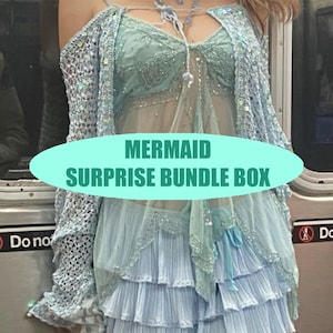 May include: A light blue mermaid-themed outfit featuring a sheer top, layered skirt, and a crocheted shawl. The text "MERMAID SURPRISE BUNDLE BOX" is displayed on a light green oval. The outfit has a delicate, ethereal appearance.