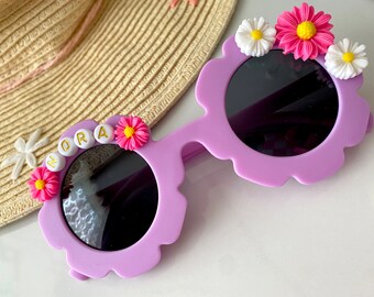 May include: Purple flower-shaped sunglasses with black lenses and white beads spelling "NORA" on one side. The sunglasses are decorated with pink and white flowers.