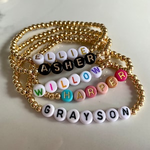 May include: Five gold beaded bracelets with different names spelled out in colorful beads. The names are Ellie, Asher, Willow, Harper, and Grayson.
