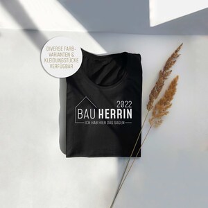 Puede incluir: Camiseta negra con texto blanco que dice "2022 Bau Herrin - Ich hab hier das sagen -" El texto está dentro de un contorno blanco de una casa.