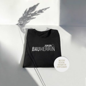 Könnte beinhalten: Schwarzes T-Shirt mit weißem Text "Junior Bauherrin" und einem kleinen Haussymbol. Ein weißer Kreis mit schwarzem Text lautet "Weitere Design-Varianten verfügbar!"