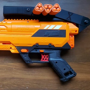 Könnte beinhalten: Orangefarbener und schwarzer Spielzeug-Blaster mit einem Magazin, das oben befestigt ist. Der Blaster hat einen schwarzen Griff und einen weißen Streifen an der Seite. Der Text "CONQUER PRO" und "POWERED BY  STROZ" ist an der Seite des Blasters aufgedruckt.
