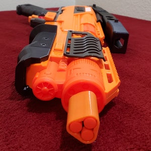 May include: An orange and black toy blaster gun with a triple-barrel design. The toy gun has a handle, trigger, and various other components. The blaster is made of plastic and appears to be a toy for children.
