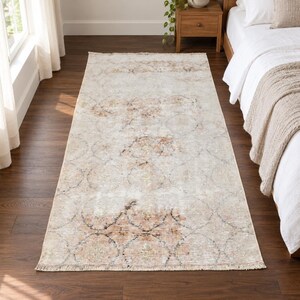 May include: A rectangular area rug featuring a faded, distressed pattern in shades of cream, beige, and muted peach. The design includes repeating circular motifs and intricate floral details, finished with short fringe along the narrow ends.