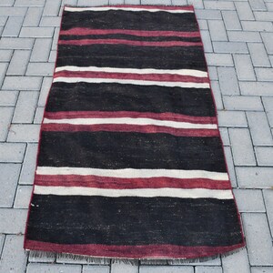 Puede incluir: Una alfombra rectangular larga con un patrón de rayas en negro, blanco y burdeos. La alfombra tiene flecos en los extremos.