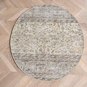 May include: A round, beige rug with an intricate, faded floral pattern. The rug has a distressed look with dark brown and gray accents. The rug is placed on a light-colored wooden floor.