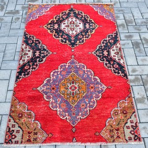 May include: A rectangular red rug with an intricate geometric pattern. The rug features a central diamond shape with orange, purple, and white details, surrounded by black and white floral motifs. The rug is placed on a grey brick surface.